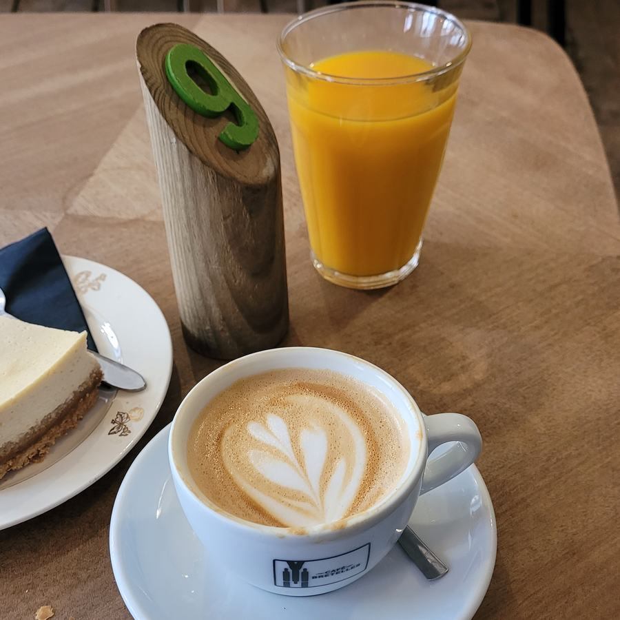Glas Orangensaft, Milchkaffee und ein Stück Käsekuchen auf einem Cafétisch